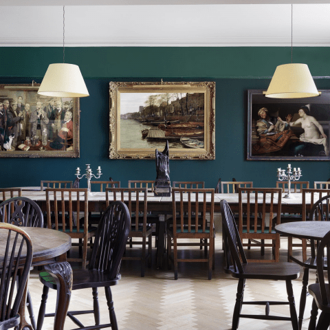 A room of tables and chairs with art work framed on the walls with lampshades hanging from the ceiling at Chelsea Arts Club